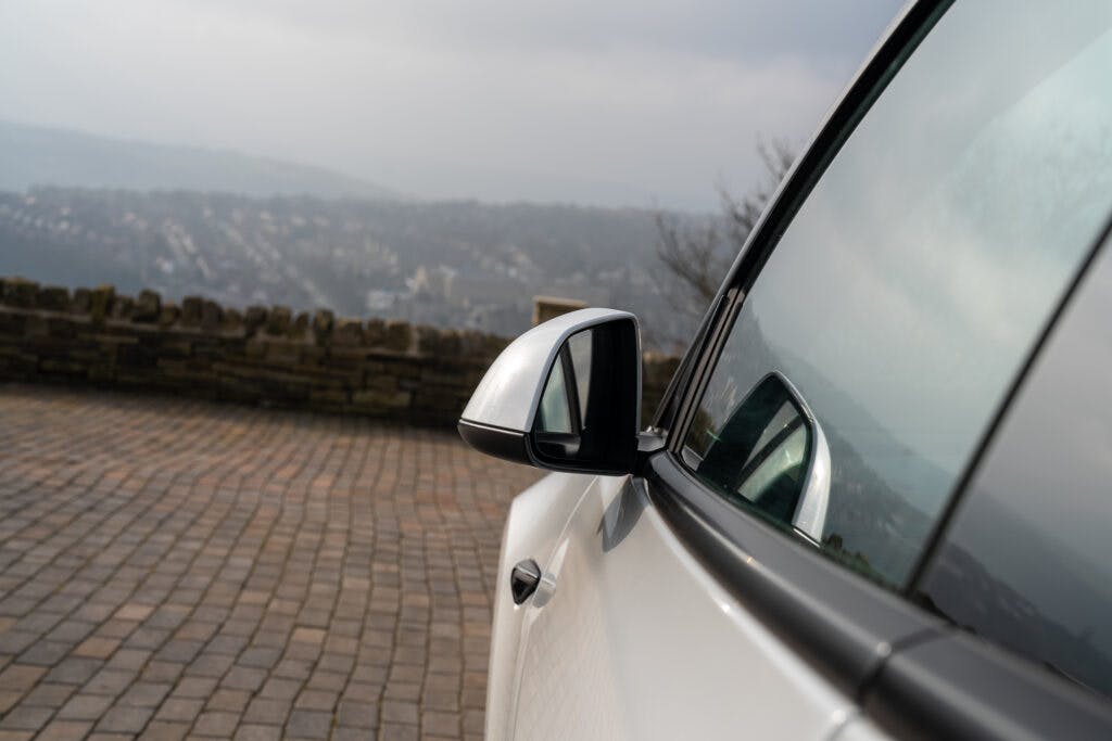 Close-up of the side mirror on a white 2021 Tesla Model 3 Long Range AWD parked on a paved surface. In the background, there's a stone wall and a distant view of a foggy landscape with blurred buildings and natural scenery.