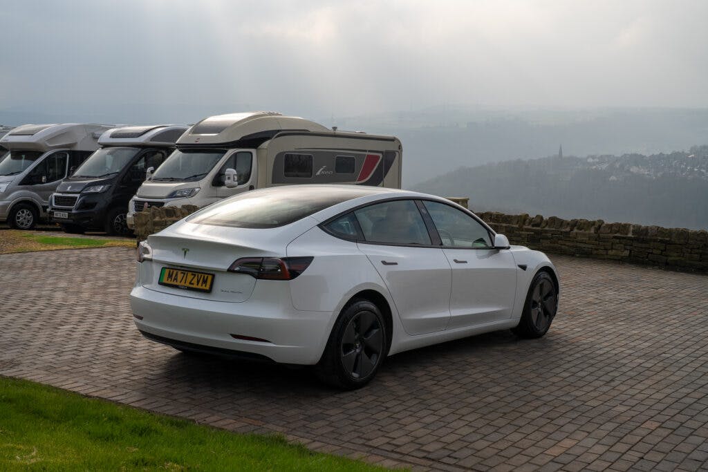 A white 2021 Tesla Model 3 Long Range AWD is parked on a paved area with a scenic backdrop of hills and a town in the distance. Several motorhomes are also parked nearby. The sky is overcast, with sunlight breaking through the clouds.