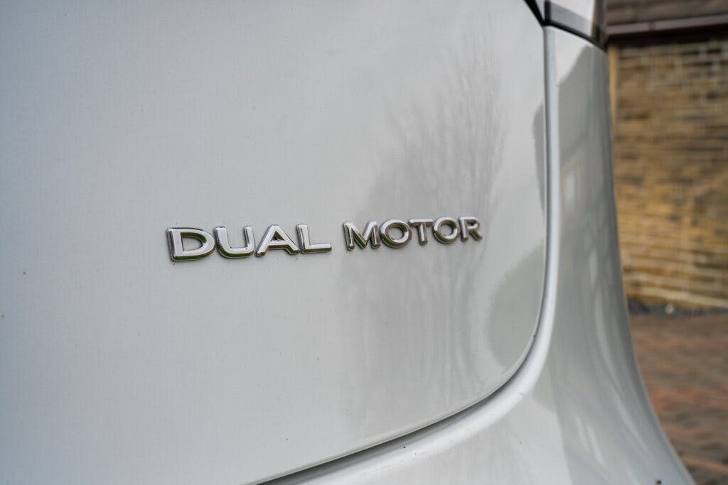 Close-up of the "DUAL MOTOR" badge on the rear of a 2021 Tesla Model 3 Long Range AWD. The background includes a blurred brick wall and driveway.