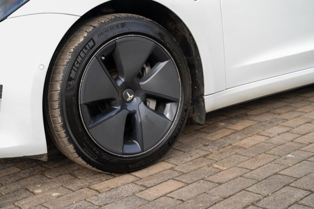 Close-up of a black car wheel with a Michelin tire on a 2021 Tesla Model 3 Long Range AWD. The car is parked on a cobblestone surface. The wheel has a distinctive design with covered spokes, and the tire appears slightly dirty, possibly from recent use.