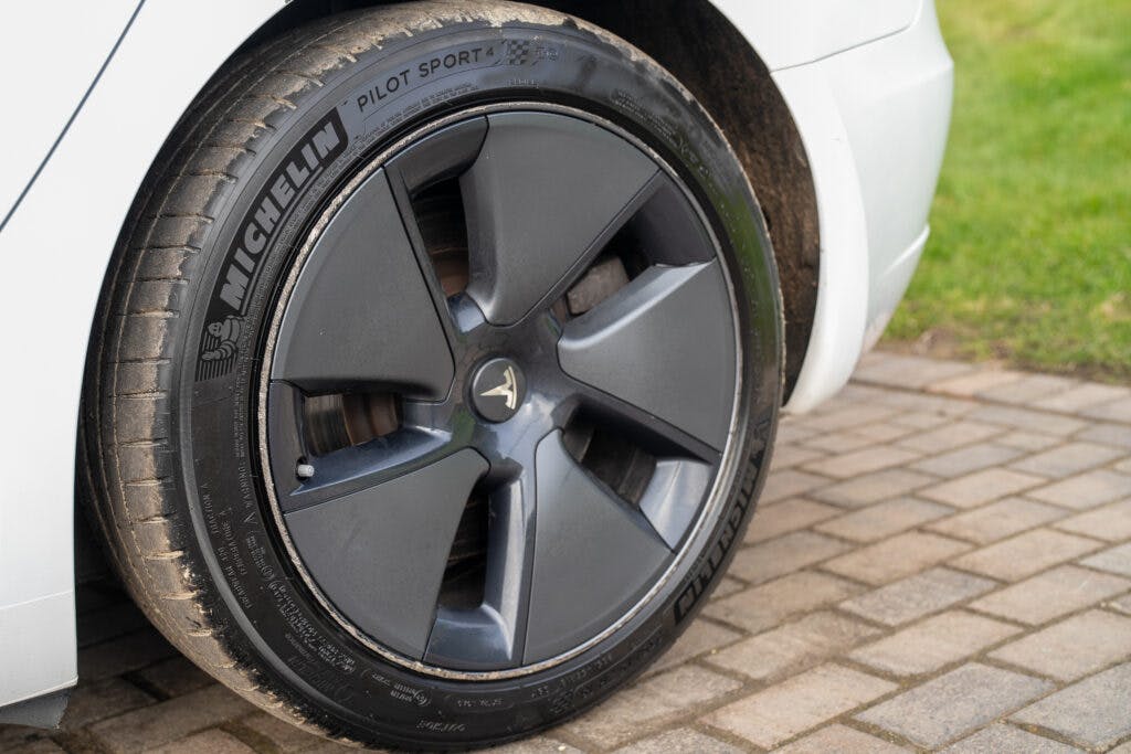 A close-up of a 2021 Tesla Model 3 Long Range AWD's front wheel, showcasing a Michelin Pilot Sport 4 tire mounted on a dark grey alloy rim with a distinctive design. The wheel is on a white car, which is parked on a paved surface with some grass visible in the background.
