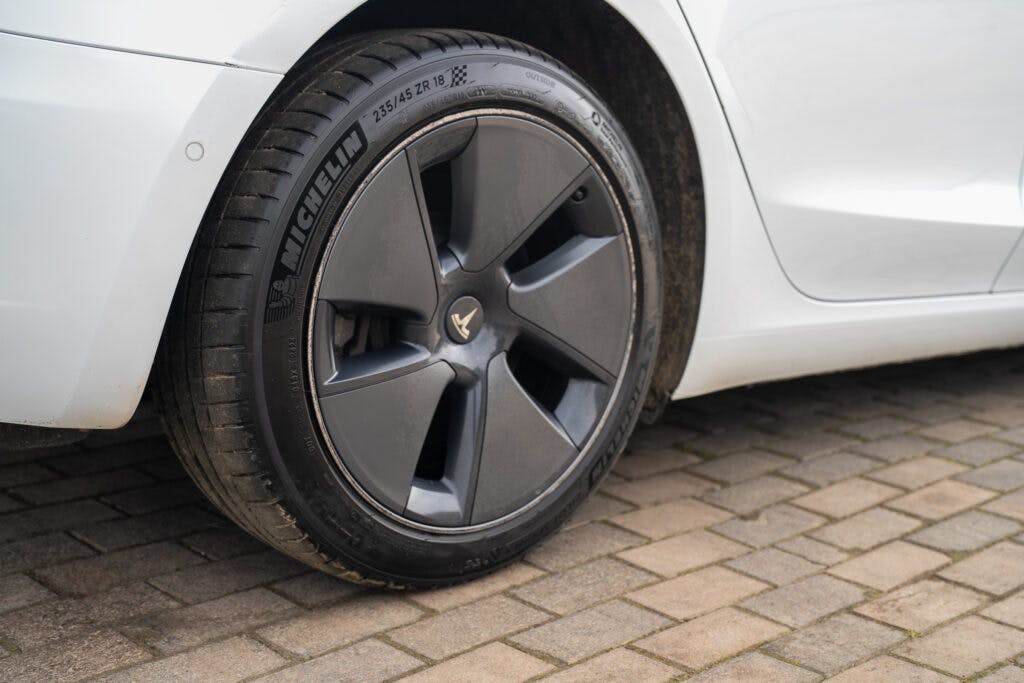 The image shows a close-up view of the rear wheel of a white 2021 Tesla Model 3 Long Range AWD. The car is equipped with Michelin tires, and the wheel has a modern five-spoke design. The vehicle is parked on a cobblestone surface.
