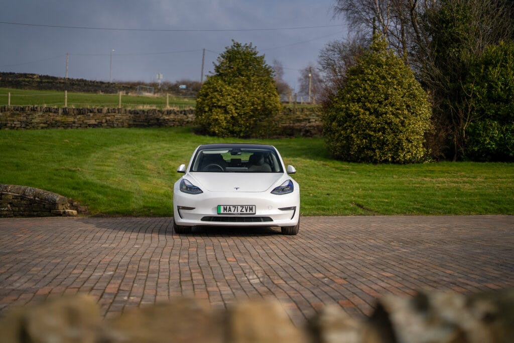 A white 2021 Tesla Model 3 Long Range AWD is parked on a brick driveway in a rural setting. The front license plate reads "NA17 VZN." There is green grass, shrubbery, and a stone wall in the background under a partly cloudy sky.