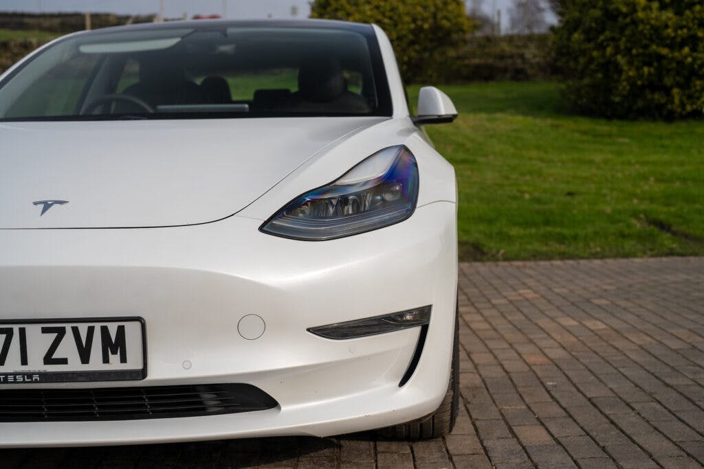 A white 2021 Tesla Model 3 Long Range AWD is parked on a paved area. The front end of the car is visible, showcasing the headlights, logo, and part of the license plate with the number "71 ZVM." There is a grassy area and a stone wall in the background.