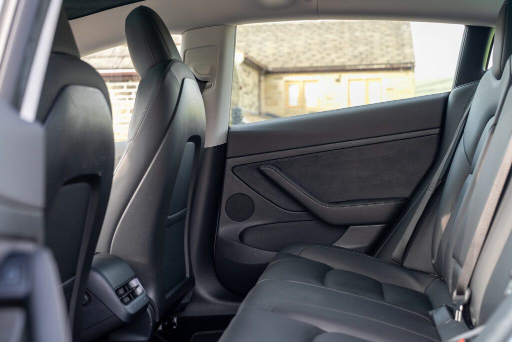 The image showcases the interior of a 2021 Tesla Model 3 Long Range AWD, focusing on the back seats. The car features sleek black leather seats and door panels. The view from the front highlights ample legroom, refined seat design, and a glimpse of the exterior through the windows.