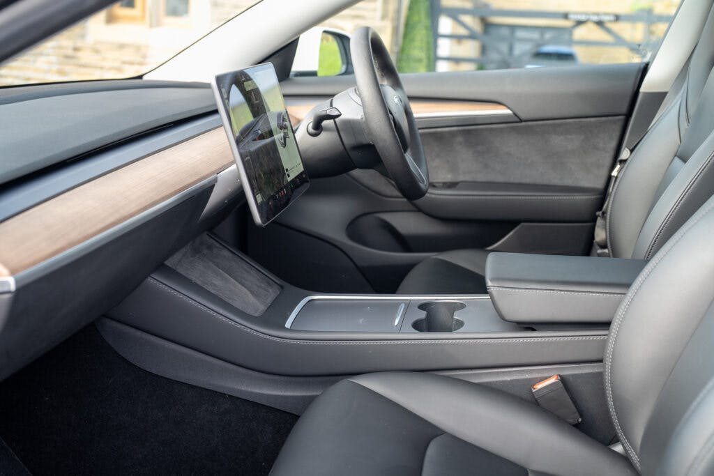 The image shows the interior of a 2021 Tesla Model 3 Long Range AWD with a minimalist design. The dashboard features a large touchscreen mounted in the center. The seating is gray with leather upholstery, and the center console has storage compartments and cup holders.