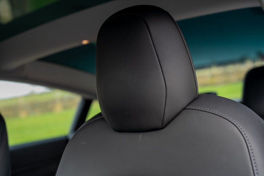 Close-up view of the back of a car seat with a black leather headrest in the sleek 2021 Tesla Model 3 Long Range AWD. The image captures the inside of the vehicle, and through the window, a grassy field and a fence add to the serene background.