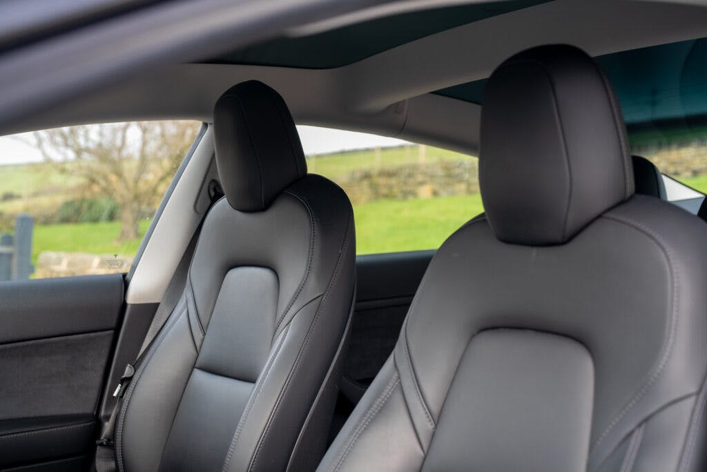 The image shows the interior of a 2021 Tesla Model 3 Long Range AWD, focusing on the two front seats. The seats are upholstered in black leather with a minimalist design. The background outside the car window reveals a green, grassy field with a stone wall.