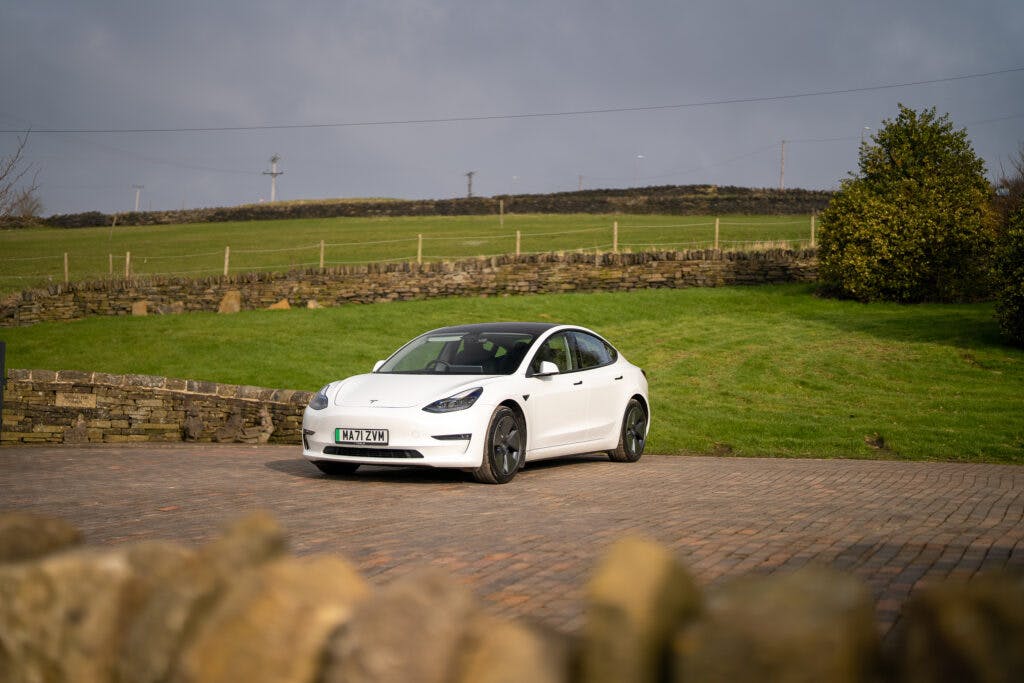 A sleek 2021 Tesla Model 3 Long Range AWD is parked on a paved area with a green field and a stone wall in the background. The sky is partly cloudy, adding to the serene rural or suburban setting. The license plate reads "MJ21 XYW.