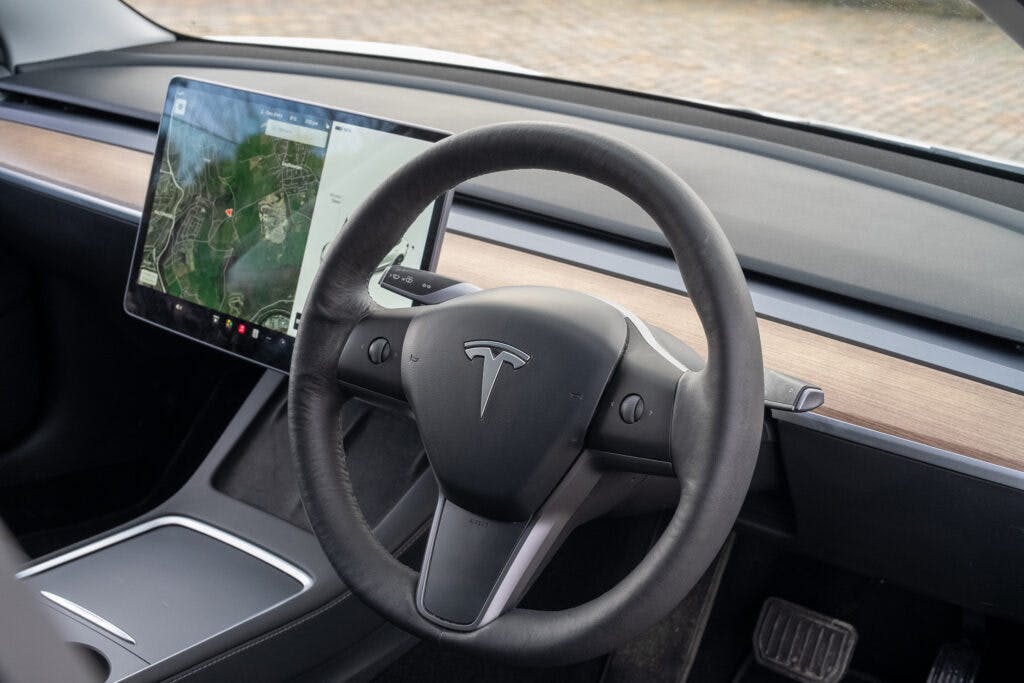 The interior of the 2021 Tesla Model 3 Long Range AWD showcases the steering wheel, dashboard, and a large touchscreen display with a map. Its minimalist design features wooden trim and black seating surfaces. The brake and accelerator pedals are also visible.