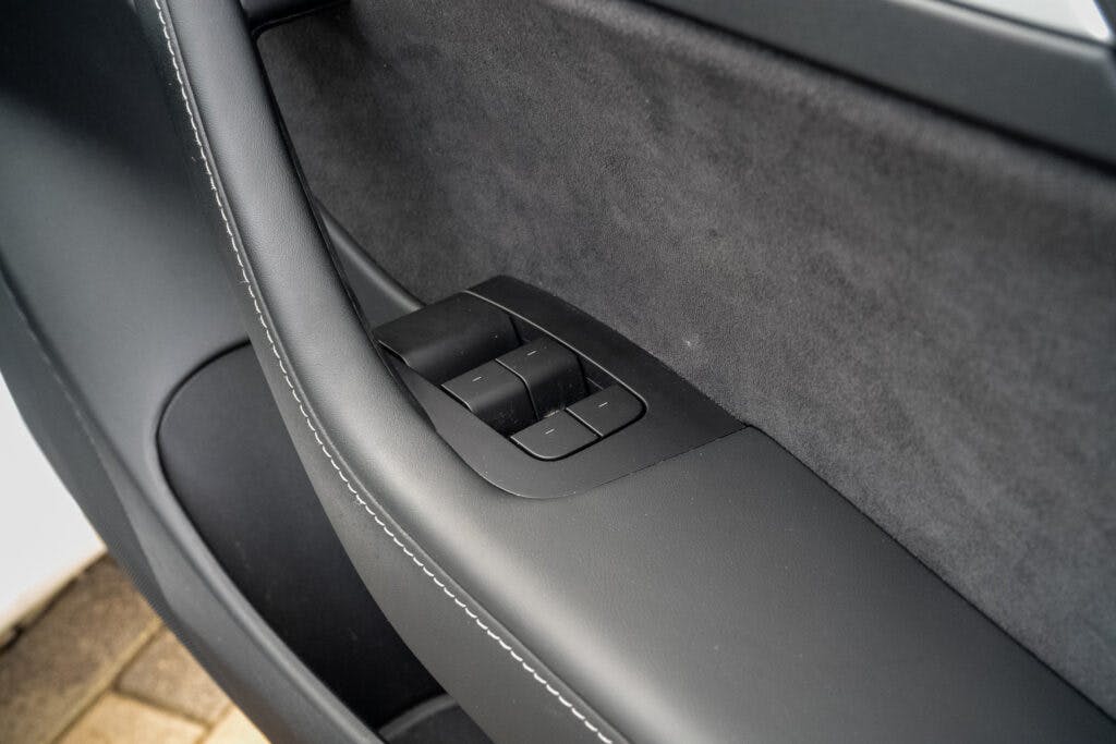 Close-up shot of a 2021 Tesla Model 3 Long Range AWD door panel featuring black electric window control buttons. The panel includes a fabric section and is outlined with white stitching. The photo highlights the sleek and modern design of the car's interior.
