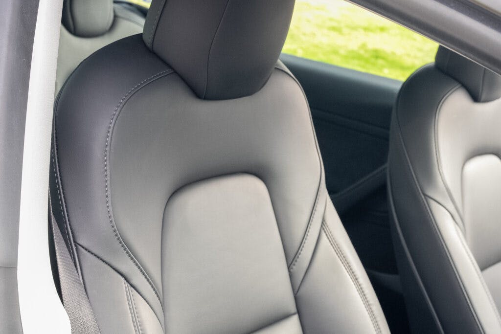 This image shows the interior of a 2021 Tesla Model 3 Long Range AWD, focusing on the sleek, black leather front seat. The seat is well-cushioned, with a headrest and stitching details. A glimpse of the seatbelt and part of the car door can also be seen.