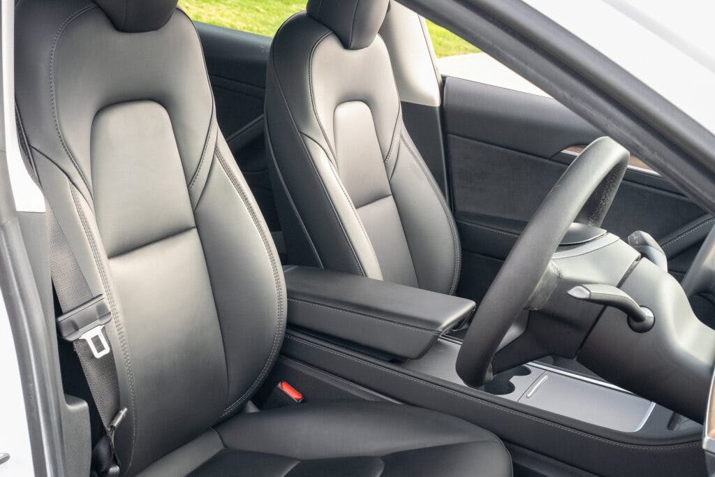 The image shows the interior of a 2021 Tesla Model 3 Long Range AWD, focusing on the front seats, steering wheel, and center console. The seats are upholstered in black leather and there is a minimalist dashboard design. The armrest and cupholders are visible in the center console.