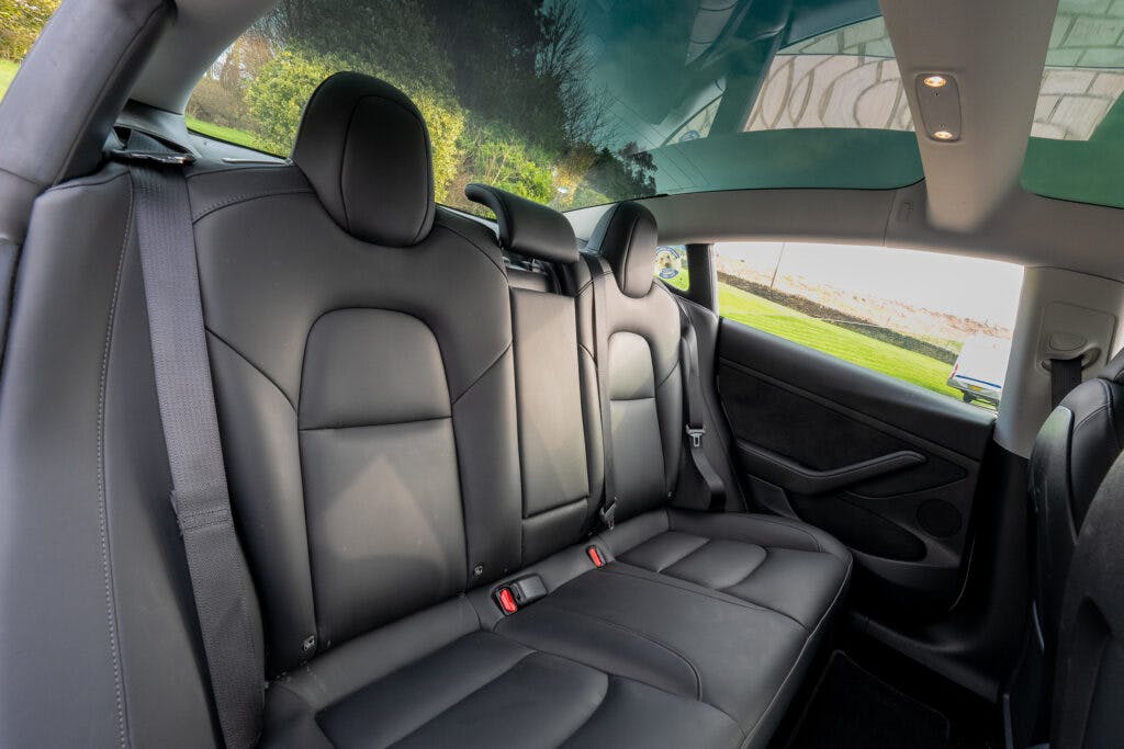 Interior view of the back seat of a 2021 Tesla Model 3 Long Range AWD with black leather upholstery, three headrests, and seat belts. The glass roof is partially visible, showing a clear sky and greenery outside.