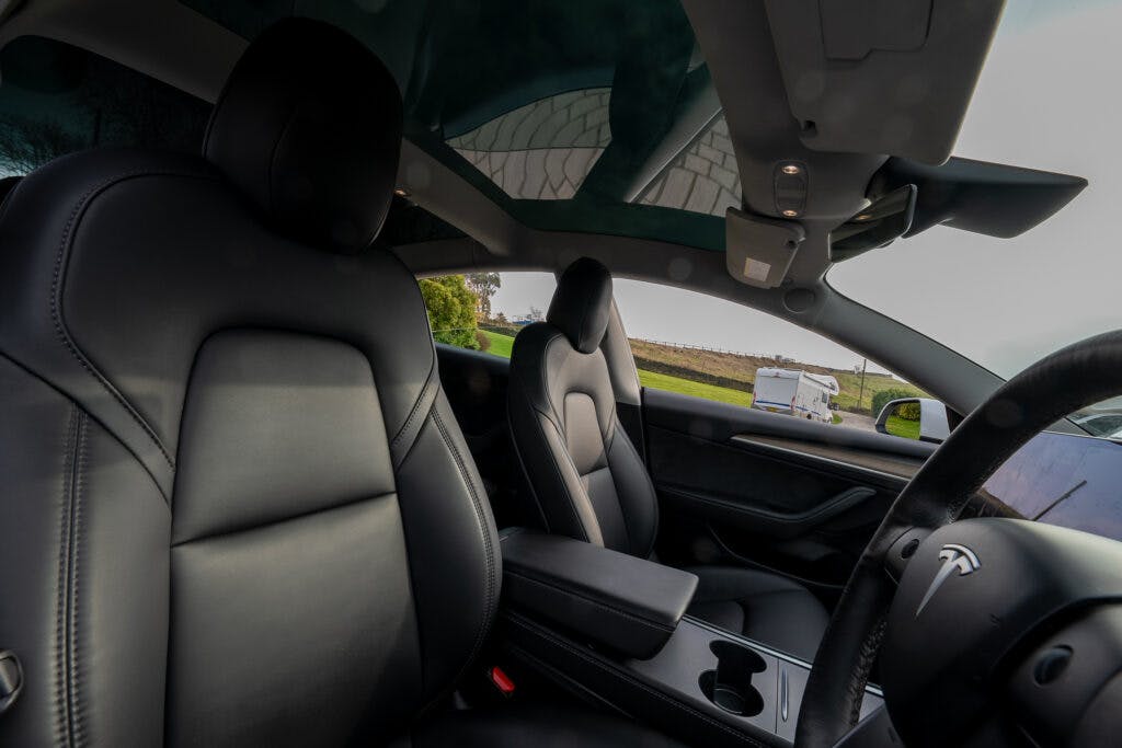 Interior view of the 2021 Tesla Model 3 Long Range AWD showcasing black leather seats, a central console with cup holders, and a panoramic sunroof. The Tesla logo is visible on the steering wheel. A scenic landscape with greenery and a road is seen through the windshield and windows.