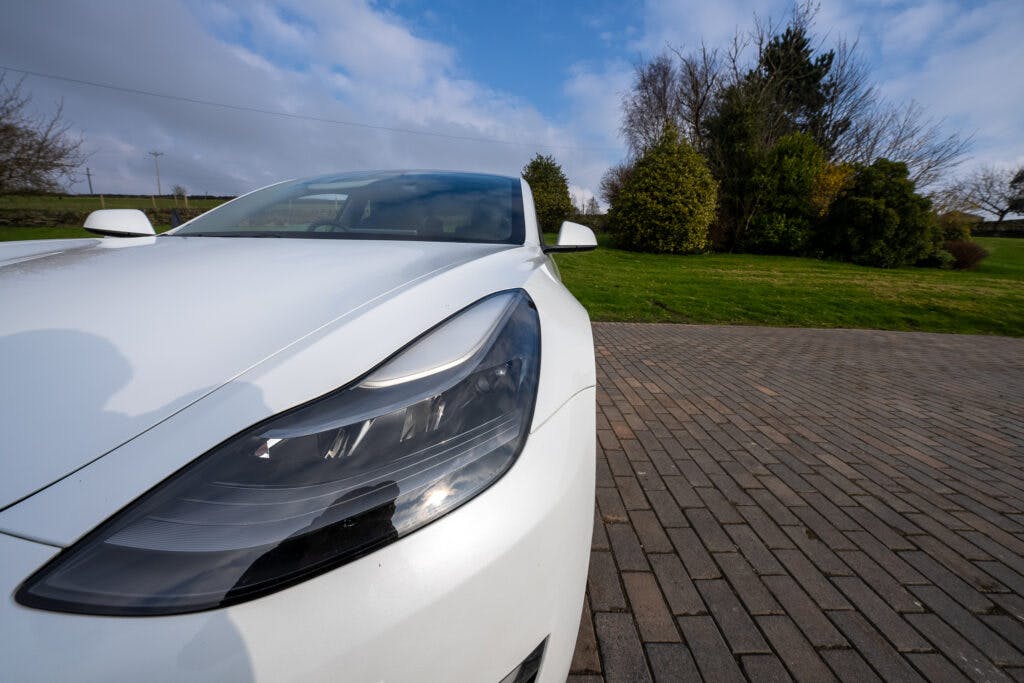 A close-up view of the front left headlight of a 2021 Tesla Model 3 Long Range AWD parked on a brick driveway. In the background, there is a green lawn, some trees, and a partly cloudy sky.