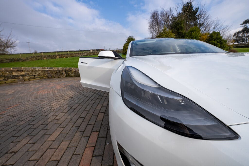 A white 2021 Tesla Model 3 Long Range AWD is parked on a paved driveway with the driver's door open. The sky is partly cloudy, and there is greenery and a low stone wall in the background.
