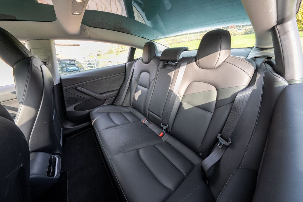 This image shows the rear interior of a 2021 Tesla Model 3 Long Range AWD with black leather seats. The seating area includes a bench that can accommodate three passengers, seatbelt fixtures, and a sunroof above. Some reflections and a person outside the car are visible through the rear window.