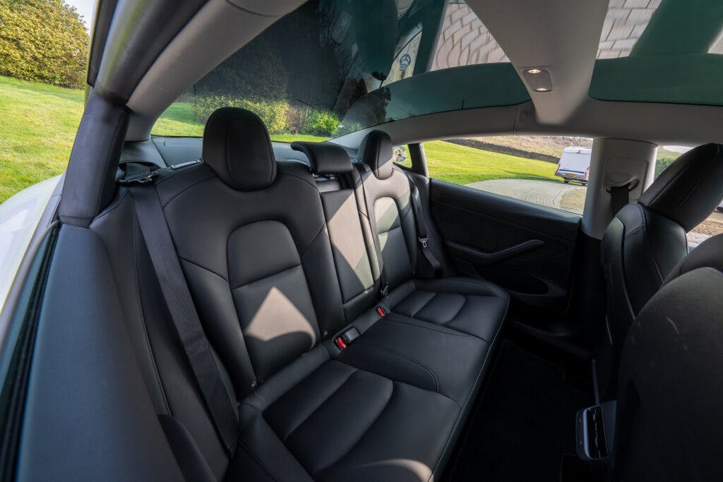 The interior of a 2021 Tesla Model 3 Long Range AWD showcasing the backseat area. The black leather seats have seat belts for three passengers. Natural light pours in through the large rear and side windows, and the car appears parked with greenery visible outside.