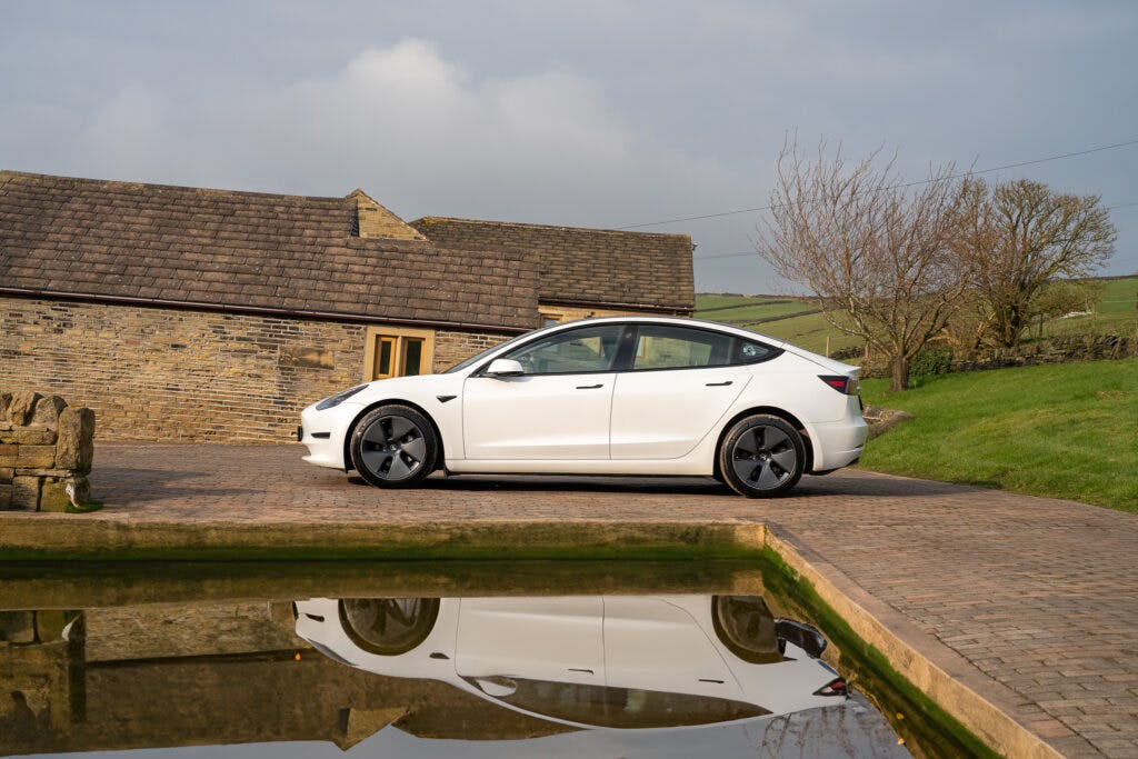 A 2021 Tesla Model 3 Long Range AWD is parked on a brick driveway next to a small pond, with its reflection visible in the water. The scene is set in a rural area with stone buildings and fields in the background under a cloudy sky.