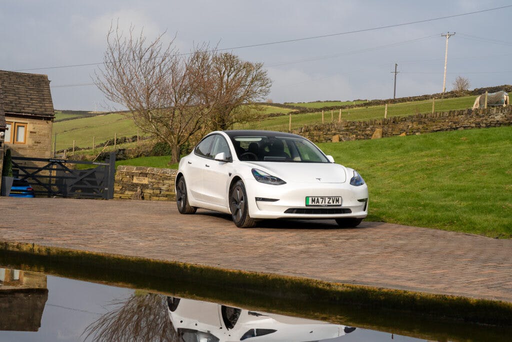 A white 2021 Tesla Model 3 Long Range AWD is parked on a stone driveway next to a rural house and a reflective pond. The backdrop shows a scenic countryside with a stone wall, green fields, and a few bare trees. The license plate reads "MA71 ZVW".