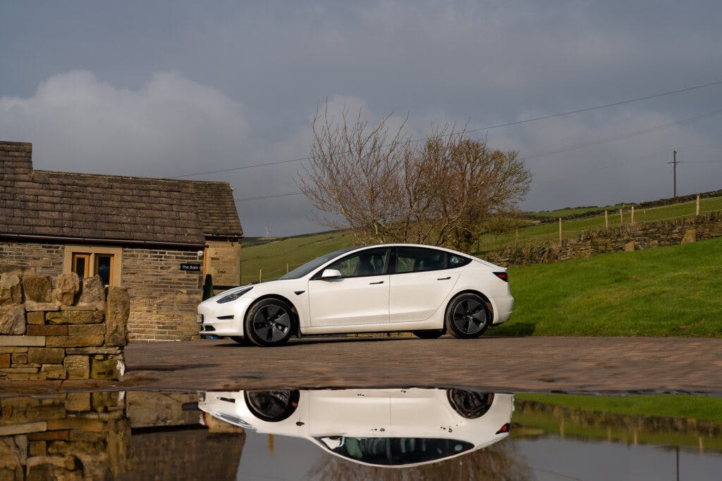 A white 2021 Tesla Model 3 Long Range AWD is parked in front of a stone building with a tiled roof. The car and the building are reflected in a puddle on the ground. The background includes grassy hills and a cloudy sky.