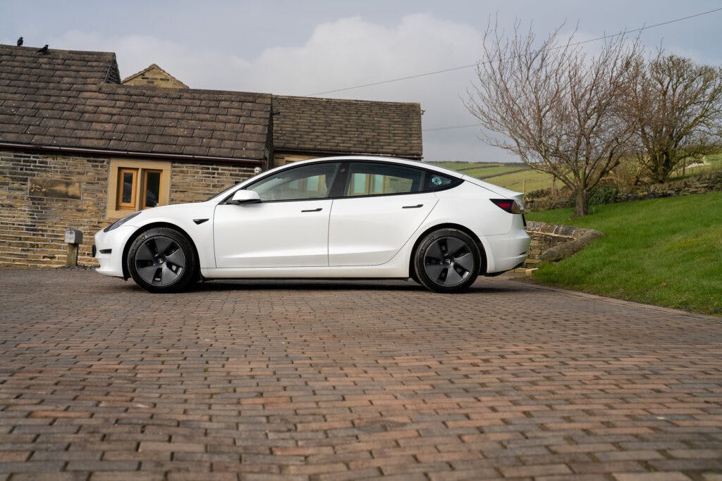 A white 2021 Tesla Model 3 Long Range AWD is parked on a cobblestone driveway in front of a stone house with a wooden door. Leafless trees and rolling hills are visible in the background. The sky is cloudy.