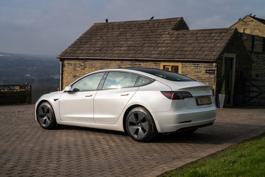 A 2021 Tesla Model 3 Long Range AWD, viewed from the rear and left side, is parked on a brick driveway. In the background, there is a stone building with a brown roof and a distant view of a landscape under a cloudy sky.
