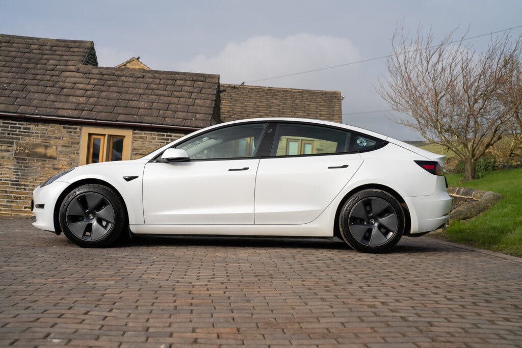 A white 2021 Tesla Model 3 Long Range AWD is parked on a paved driveway in front of a brick house. The house has a sloped, tiled roof, and there are leafless trees visible in the background. The sky is cloudy.