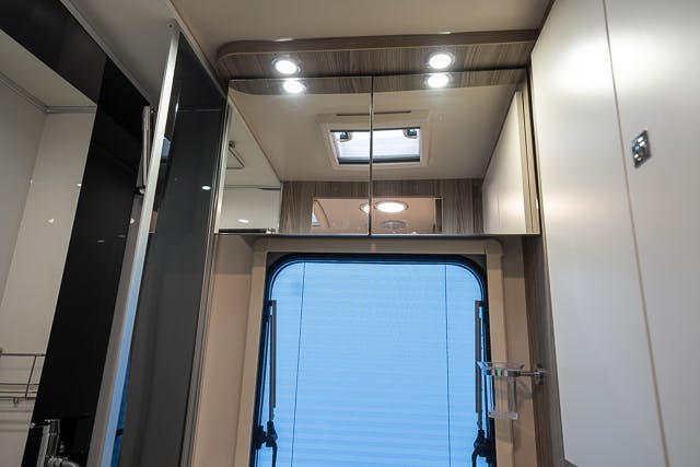 An interior view of the 2019 Benimar Tessoro T486 bathroom reveals a modern design with a skylight, mirror, recessed lighting, and a window with a roller blind. The predominantly white walls are complemented by a hint of a shower space on the left side.