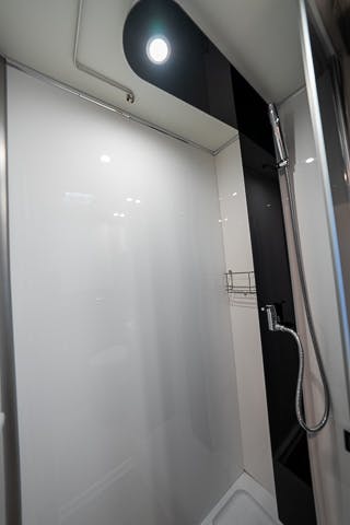 A modern shower stall in the 2019 Benimar Tessoro T486 features a minimalist design with clean, white walls and a sleek, black panel. It includes a handheld showerhead attached to a flexible hose and a metal rack for storage. An overhead light illuminates the shower space.