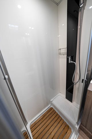 A modern shower in the 2019 Benimar Tessoro T486, featuring a glass door, wooden slatted floor mat, built-in metal soap rack, and handheld showerhead attached to a vertical rail. The wall and floor are light-colored with a dark accent panel on one side.