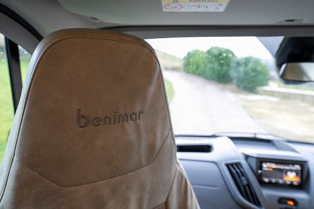 The image shows the interior of a 2019 Benimar Tessoro T486, focusing on the headrest of the driver's seat, which has "Benimar" embroidered on it. The windshield and dashboard, including a navigation screen, are visible. There's a road and greenery seen through the windshield.