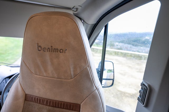 The image shows a close-up view of the driver's seat inside a 2019 Benimar Tessoro T486, with "benimar" embroidered on the headrest. The window next to the seat is slightly open, revealing an outside view with rain droplets on the glass.