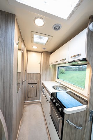 A narrow RV kitchen in the 2019 Benimar Tessoro T486 features wood-paneled walls and white cabinets. It includes a stove, oven, sink, and countertop on the right side. A window above the counter and a door at the end of the hallway enhance its practicality, with ceiling lights providing ample illumination.
