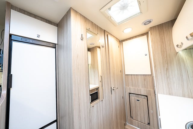 A modern RV interior featuring light wood paneling in the 2019 Benimar Tessoro T486. The image shows a white refrigerator, several storage cabinets, a small microwave, and a ceiling vent. The space is bright and well-lit with sleek, minimalist design elements.
