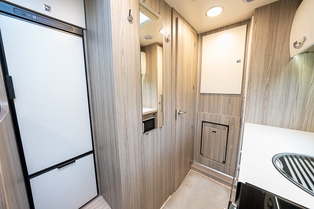 Interior of the 2019 Benimar Tessoro T486 RV kitchen and hallway area, featuring light wood cabinetry, a narrow hallway with a refrigerator on the left, storage cabinets, and a compact countertop with a sink on the right. Ceiling lights provide ample brightness.