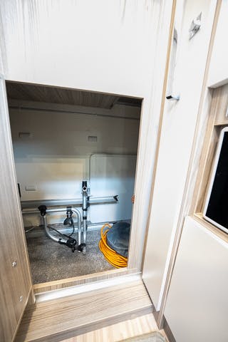 A compact storage space in the 2019 Benimar Tessoro T486 features a metal bicycle rack and a coiled orange extension cord. The space is enclosed by light wooden panels, with part of a wall-mounted control panel visible on the right side.