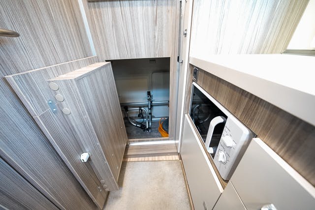 A view of a modern, compact kitchen area inside a 2019 Benimar Tessoro T486. The image shows wooden cabinetry, a microwave, and an open compartment revealing storage space and plumbing pipes. The light-colored flooring complements the wooden and white surfaces perfectly.