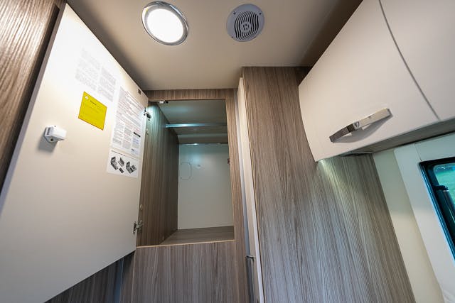 Interior of a light wood-finished cabinet in a 2019 Benimar Tessoro T486 with an open door, revealing an empty shelf and some documents attached to the inner side of the door. A circular light and air vent are mounted on the ceiling above. Another closed cabinet is visible on the right.