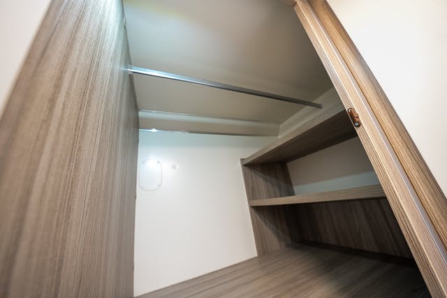 Interior view of an empty wooden closet inside the 2019 Benimar Tessoro T486, featuring two built-in shelves on the right, a metal hanging rod, and beige walls. The closet has a light-colored floor and a door that is partially open.