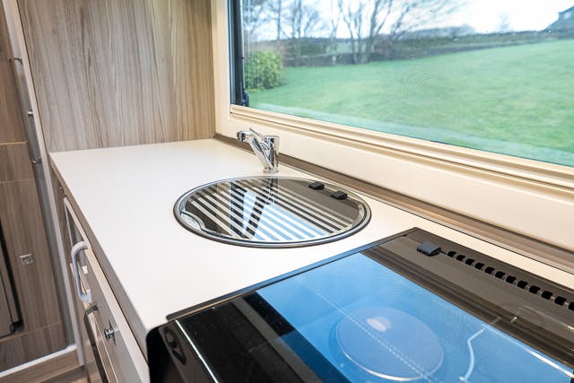 A modern kitchen area in the 2019 Benimar Tessoro T486 features a round stainless steel sink and faucet on a white countertop. Next to the sink is a black electric stove. A large window above the countertop offers a view of a grassy outdoor area, and light wood cabinets complete the look.