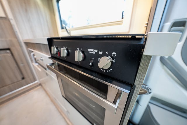 A close-up view of a black Thetford Triplex kitchen stove inside the modern, compact kitchen area of a 2019 Benimar Tessoro T486. The stove features three control knobs, a slim oven door beneath, and is set against a light wood and silver-toned interior backdrop.