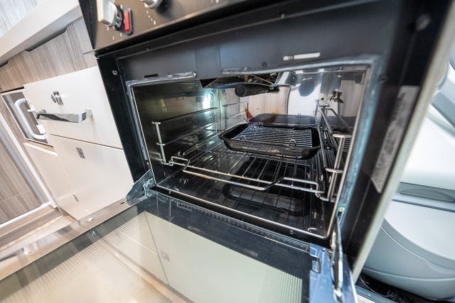 An open oven in a modern kitchen is shown with its door pulled down, reminiscent of the sleek design found in a 2019 Benimar Tessoro T486. The interior is empty except for metal racks. The surrounding area includes cabinetry, drawers, and a microwave on the left, with light reflecting off the oven's interior surfaces.