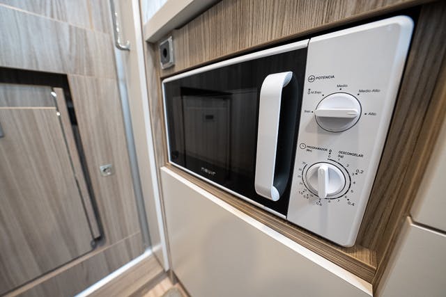 A close-up of a microwave oven built into a wooden kitchen cabinet in the 2019 Benimar Tessoro T486. The microwave features a white handle and two control knobs for power and timer settings, seamlessly blending with the modern wood finish of the surrounding cabinetry.