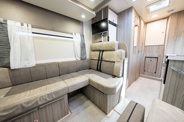 Inside the 2019 Benimar Tessoro T486, you'll find a cozy seating area with an L-shaped couch and large windows adorned with curtains. The modern space features light wood finishes, a small wall-mounted television, and ample overhead storage. A corridor leads to other well-appointed compartments in the RV.