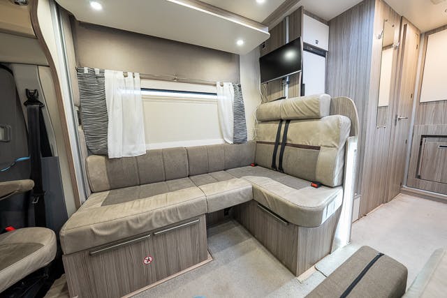 The interior of the 2019 Benimar Tessoro T486 RV features a seating area with beige cushions and a mounted TV. The space includes grey and white curtains on the window and wood-paneled walls. The RV's layout is compact, utilizing the available space efficiently.