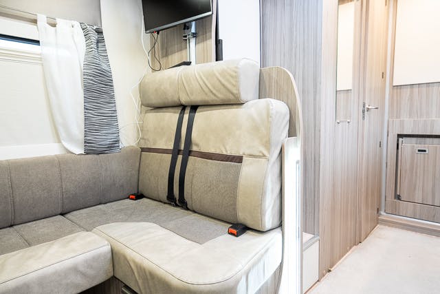 Interior of the 2019 Benimar Tessoro T486 featuring a beige cushioned bench seat with seat belts. The area is well-lit with natural light from a window with patterned curtains. A wall-mounted TV and built-in cabinets are visible in the background.