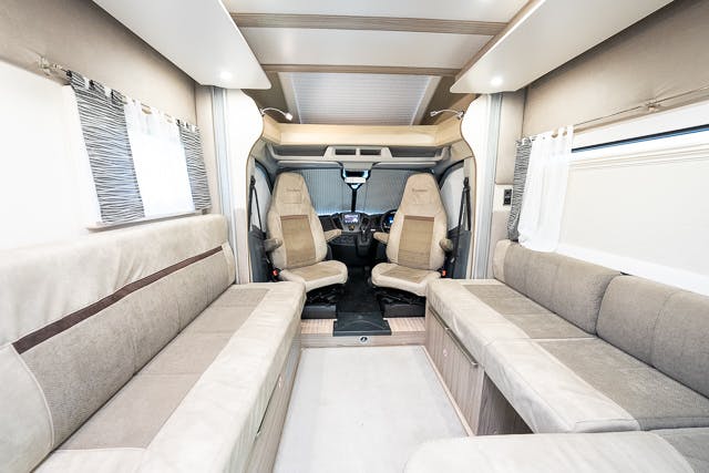 Interior of the 2019 Benimar Tessoro T486 featuring beige seating areas on both sides, a light-colored floor, and driver's and passenger's seats at the front. The windows are covered with partially drawn curtains, and there is a small step leading to the driver's area.