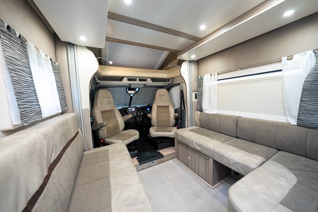 Interior of the 2019 Benimar Tessoro T486 showing a spacious seating area with light-colored upholstery. The front driving cabin has two seats, and the back area features an L-shaped couch and a table. Ambient lighting and windows with patterned and white curtains are visible.